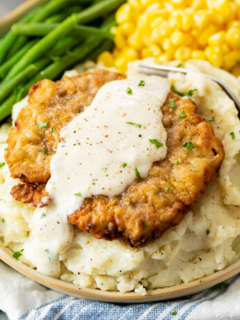 Crispy Chicken Fried Steak on a bed of mashed potatoes with white gravy on top and vegetables in the back.
