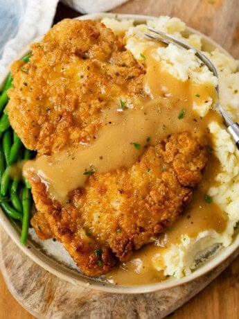 A plate with Country Fried Chicken over mashed potatoes with green beans and gravy.
