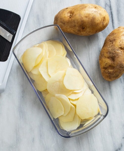 Thinly sliced potatoes in a mandoline with russet potatoes on the side.