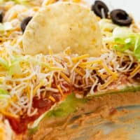 A tortilla chip on top of 7 layer dip in a baking dish.