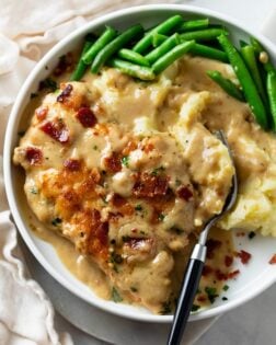 A white plate with Smothered Chicken next to mashed potatoes with gravy and green beans.