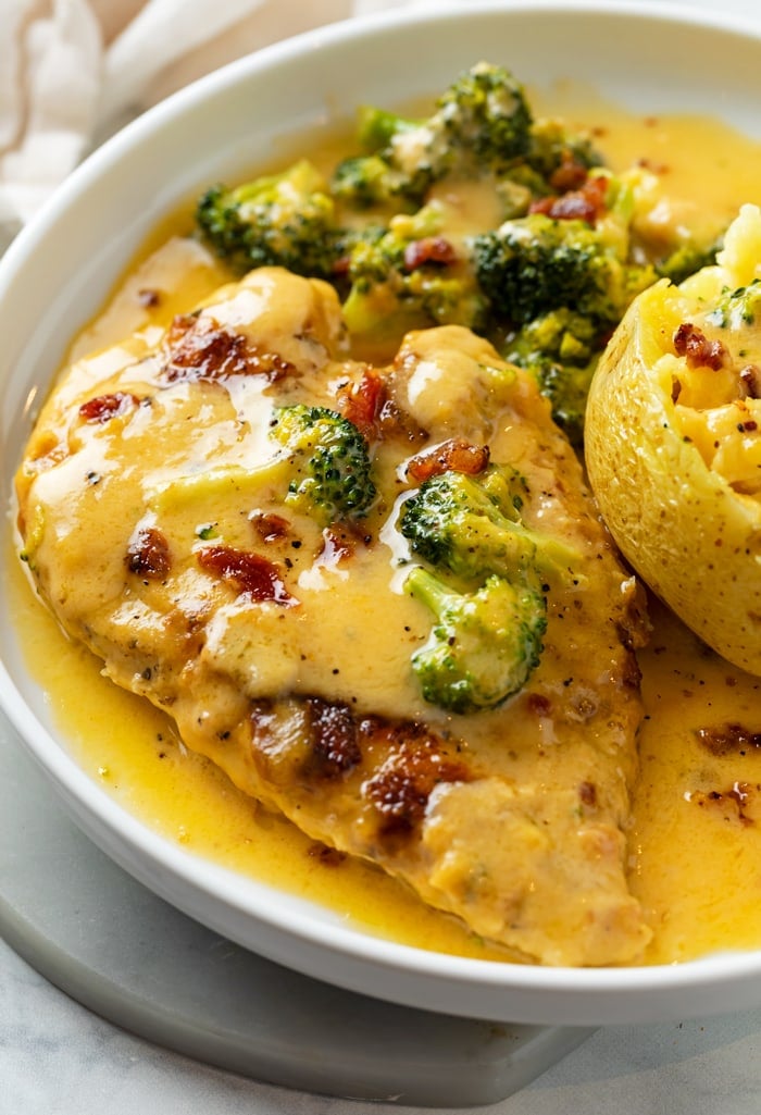A white plate with Cheese Chicken with bacon on top with broccoli and a baked potato.