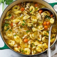 A soup pot filled with Chicken Tortellini Soup with vegetables, seasoning, and fresh parsley.