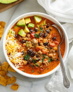 A white bowl filled with taco soup garnished with diced avocado and shredded cheese.