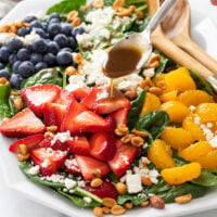 A spinach salad on a white platter with Balsamic Vinaigrette being drizzled on top.