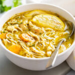 Lemon Chicken Soup in a white bowl with fresh lemon slices and a spoon on the side.