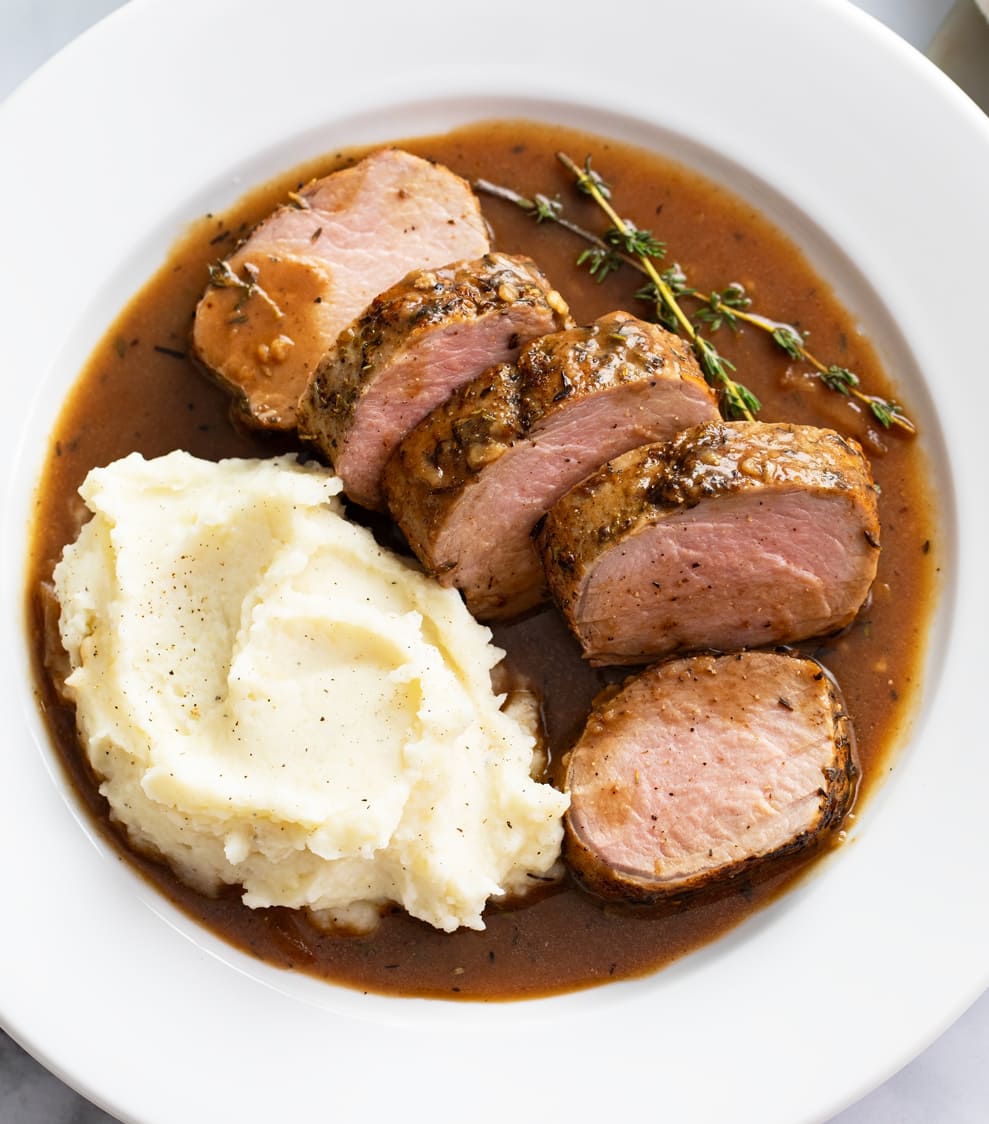 Roasted Pork Tenderloin on a white plate with pan juice, mashed potatoes, and fresh thyme.