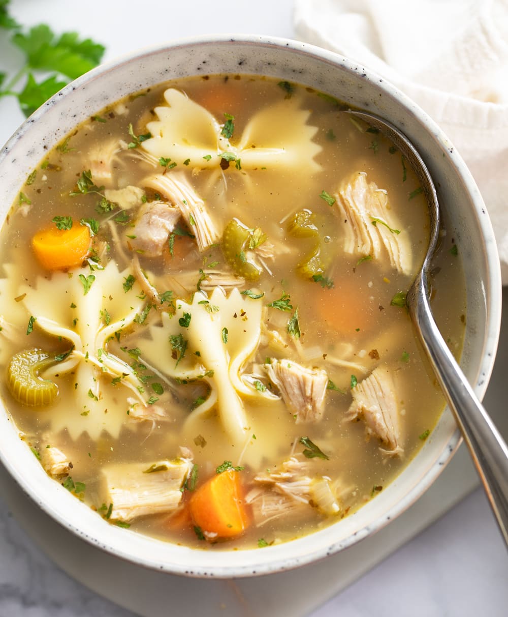 A bowl of Turkey Soup with vegetables and bowtie pasta with a spoon on the side.