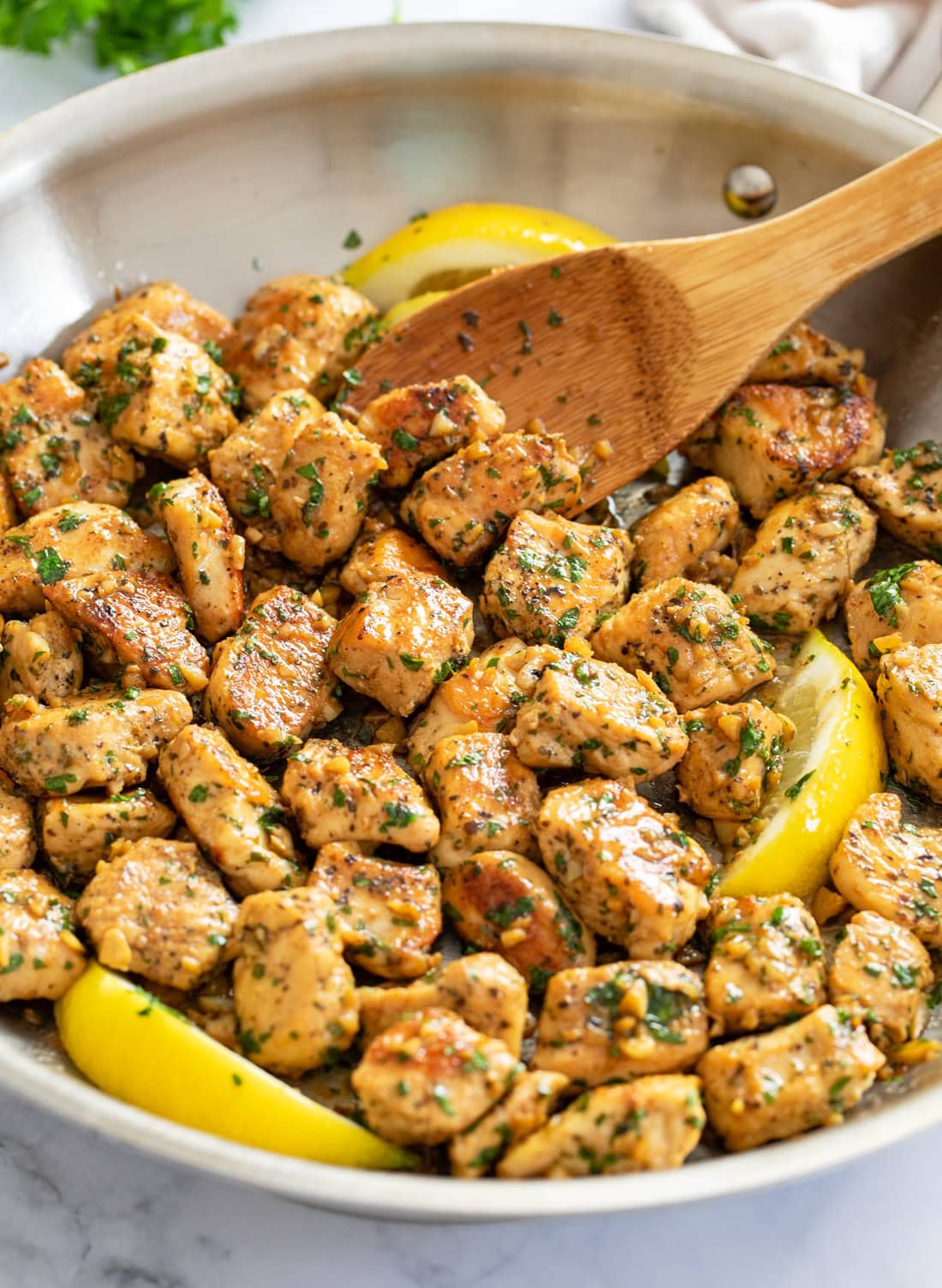 Chicken Bites in a skillet in a butter garlic sauce with lemon slices and a wooden spoon.