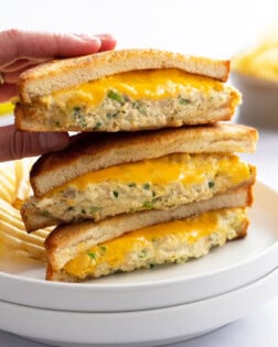 A hand taking the top Tuna Melt from a stack of 3 on a white plate with melted cheese.