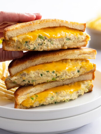 A hand taking the top Tuna Melt from a stack of 3 on a white plate with melted cheese.
