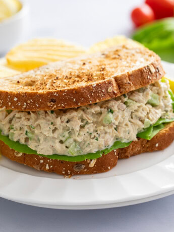 A Tuna Salad Sandwich with toasted bread on a white plate with a pickle and chips in the background.
