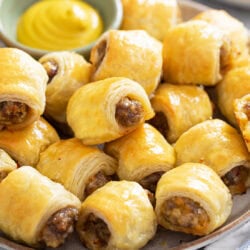 A plate of Sausage Rolls stacked up with mustard in the back for dipping.