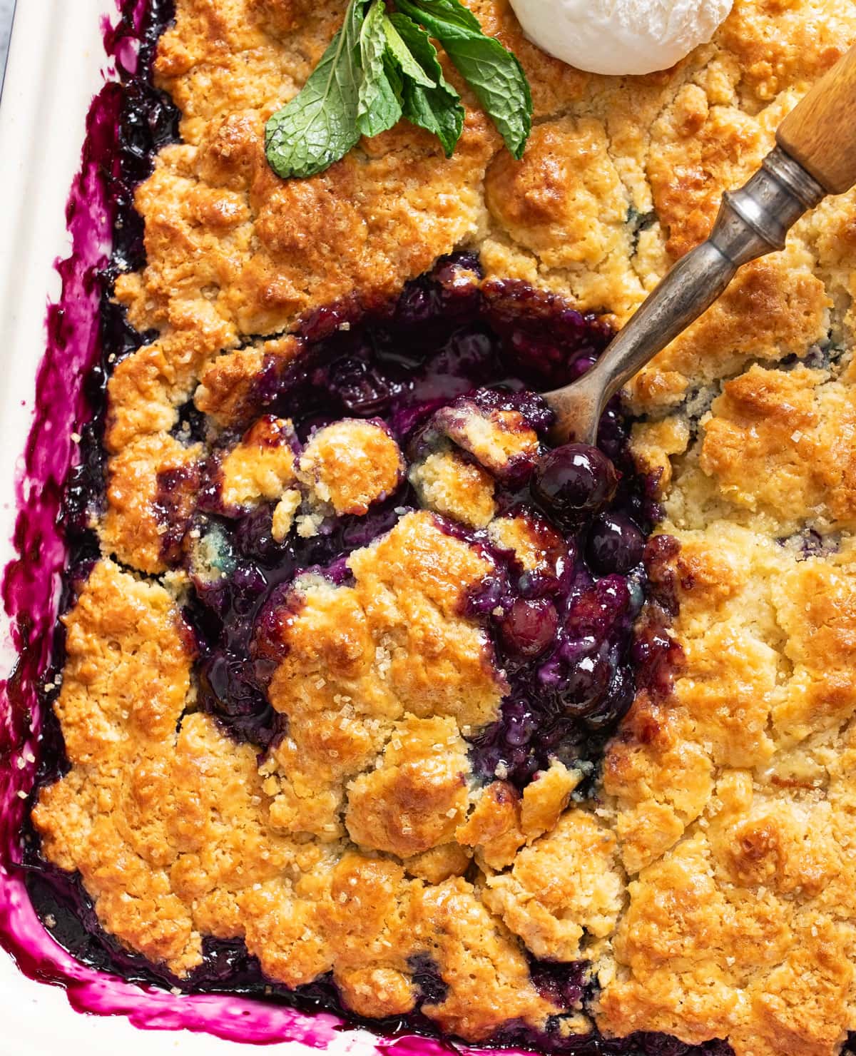 A spoon scooping Blueberry Cobbler out of a casserole dish.