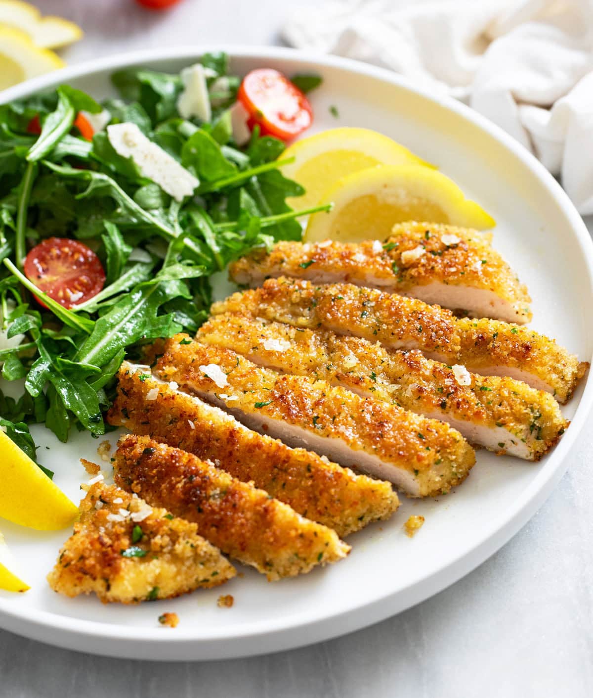 A side view of Chicken Milanese cut into slices on a white plate with lemon wedges and arugula.