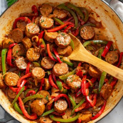 Sausage and Peppers in a skillet with a pan sauce and a wooden spoon in the middle scooping some up.