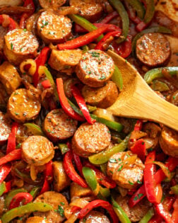 A wooden spoon scooping up sausage and peppers from a skillet.