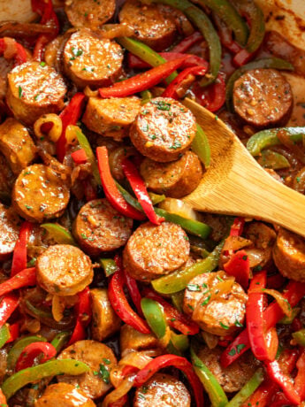 A wooden spoon scooping up sausage and peppers from a skillet.
