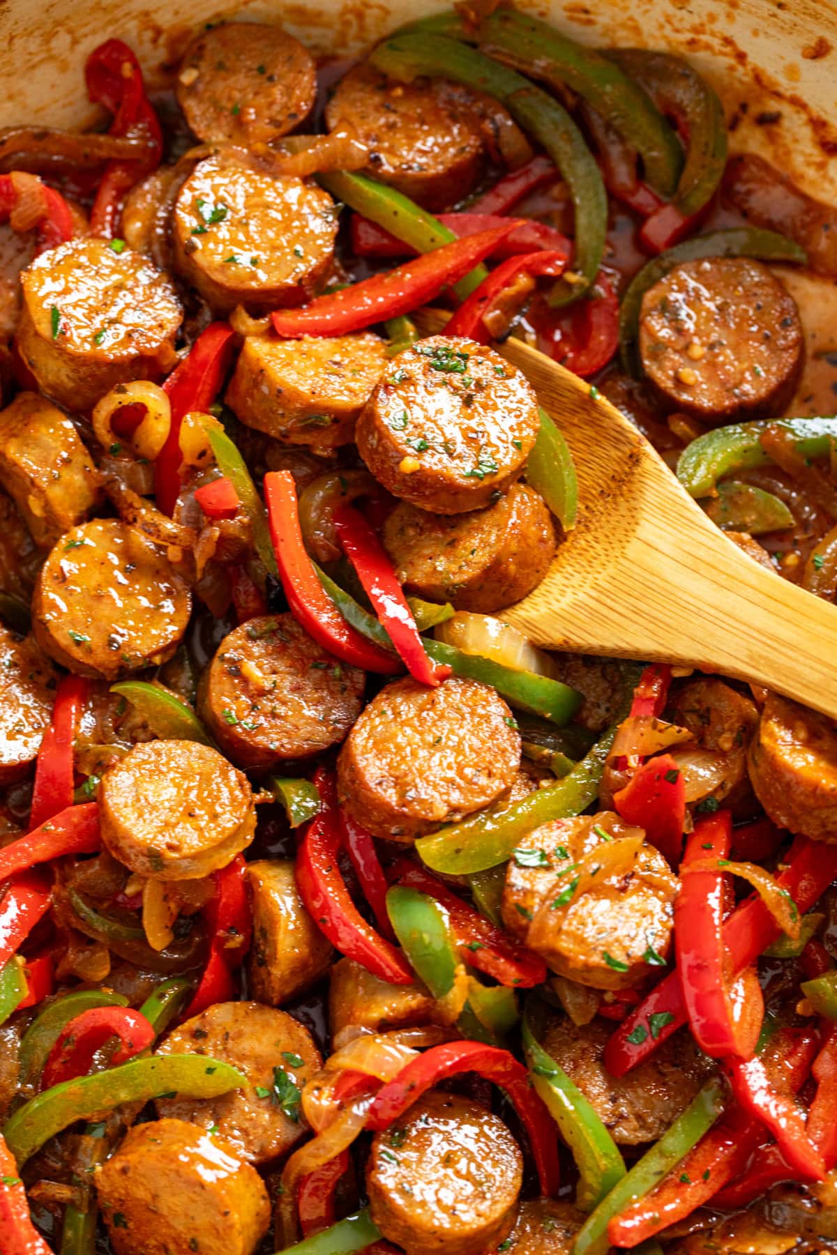 A wooden spoon scooping up sausage and peppers from a skillet.