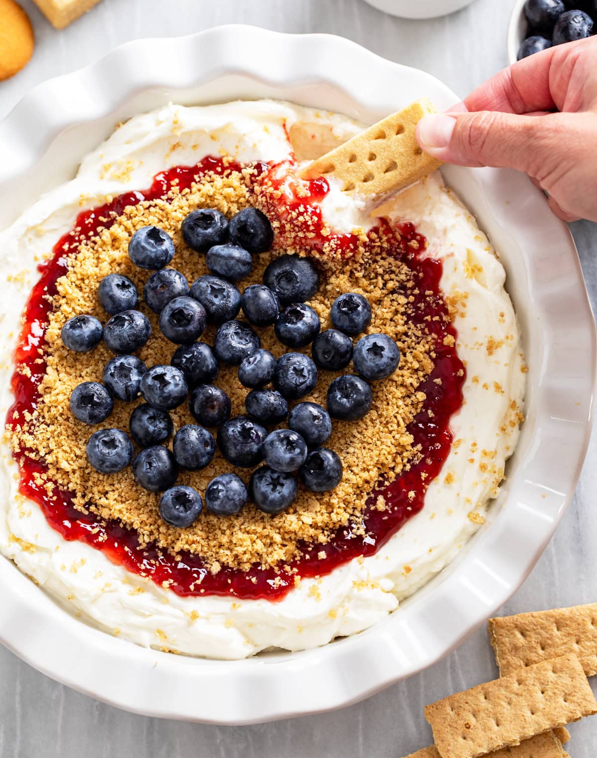 Cheesecake Dip in a pie pan with strawberry jam, graham cracker crumbs, and blueberries on top.