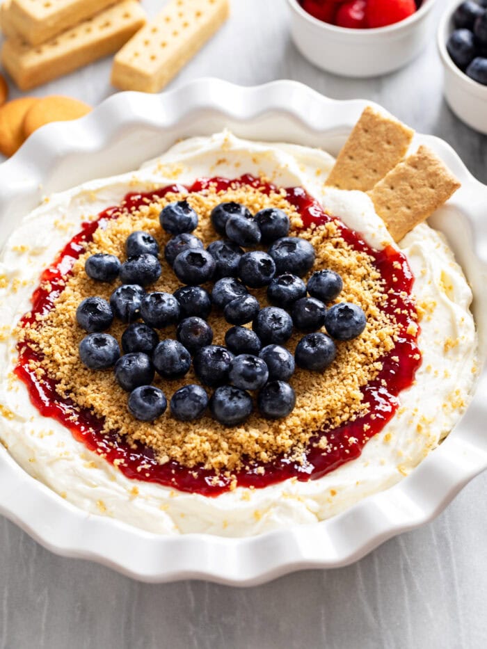 Side view of Cheesecake Dip in a pie pan topped with red jam, buttery graham cracker crumbs, and fresh blueberries.