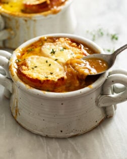 French Onion Soup in a crock with melted cheese and bread on top and a spoon scooping up the broth.